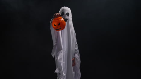 child dressed up in ghost costume trick or treating at halloween holding pumpkin shaped jack o'lantern bucket against black background