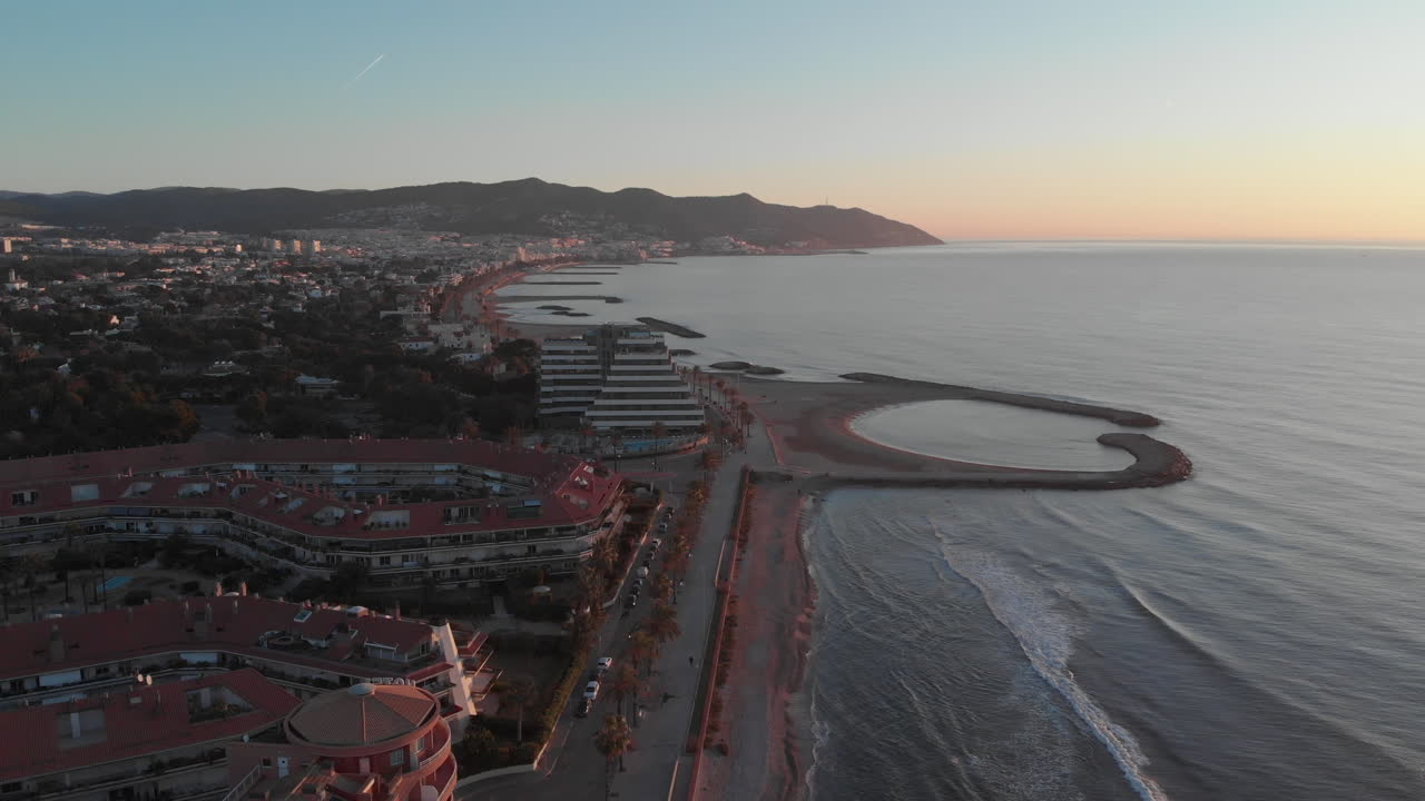 Drone flying over touristy city on coastline with mountain range standing on horizon and glowing in rising morning sun light
