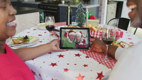 una pareja afroamericana sonriente usando una tableta para una videollamada de navidad con una pareja feliz en la pantalla