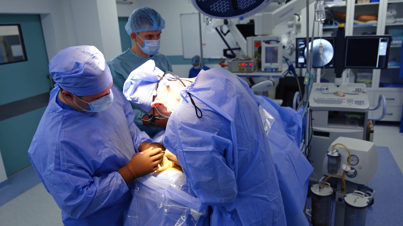 Un medico uomo si è chinato sul paziente, applicando con sforzo uno strumento chirurgico. Inquadratura dall'alto sull'équipe medica che lavora in sala operatoria.