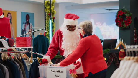 retail assistant dressed as santa claus collecting unneeded clothes from clients in donation box, giving them as present to those in need during christmas season, spreading holiday cheer