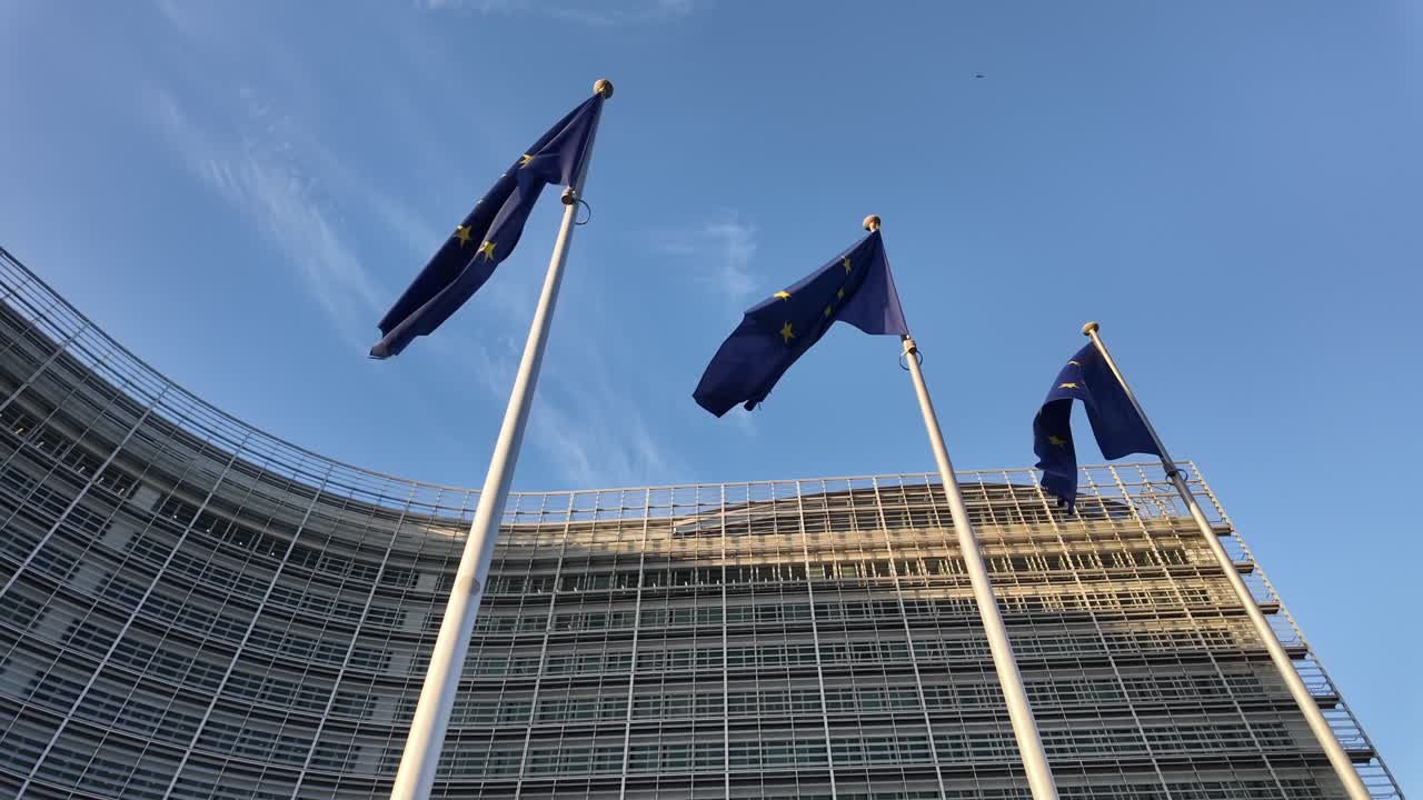 Three European Union flags flutter in the wind outside the Berlaymont building in Brussels, captured from a low angle with clear blue sky and curved glass facade in the background.