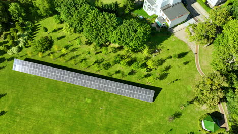 a wider aerial view of a solar panel installation on a green field, with surrounding trees and nearby buildings visible in the distance