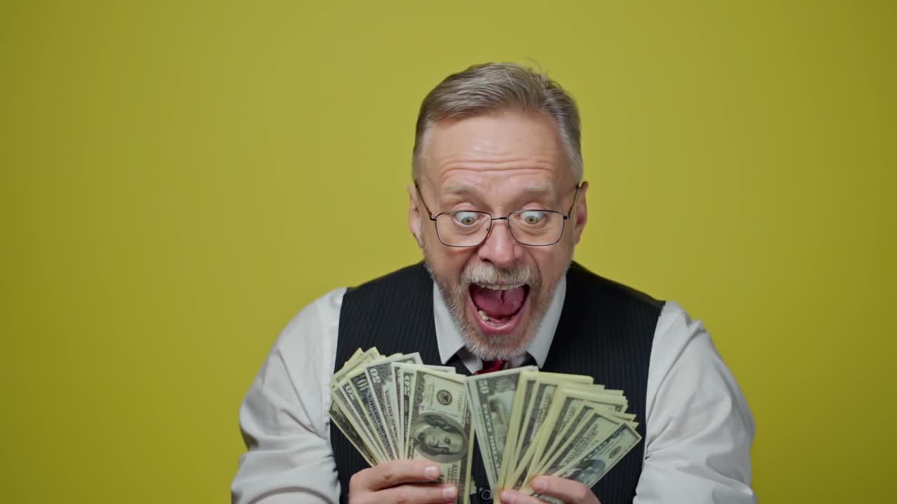 Happy senior man holding many dollar banknotes. Excited old businessman enjoys money in his hands standing alone in yellow studio.