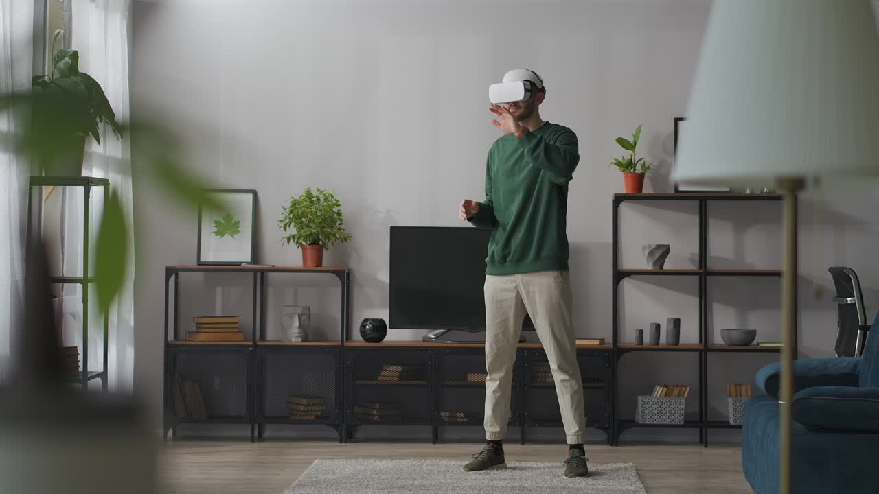 joven está usando una pantalla montada en la cabeza de pie en la sala de estar y deslizando la pantalla virtual con las manos, de longitud completa
