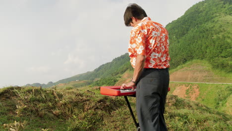 Camera-filming-man-from-behind-while-he-plays-the-piano-from-the-top-of-a-hilltop,-Vietnam