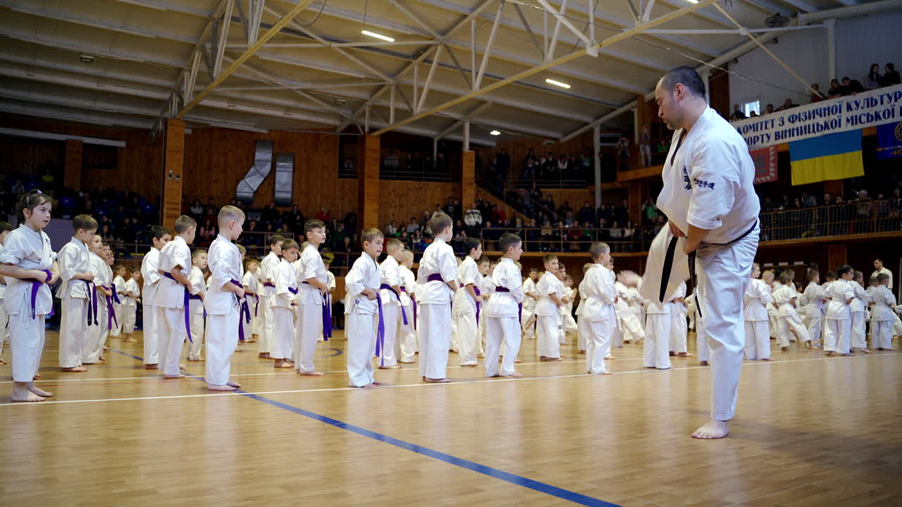 Male karate coach shows the moves with legs to his numerous trainees. Young sportsmen repeat the exercise after the trainer.