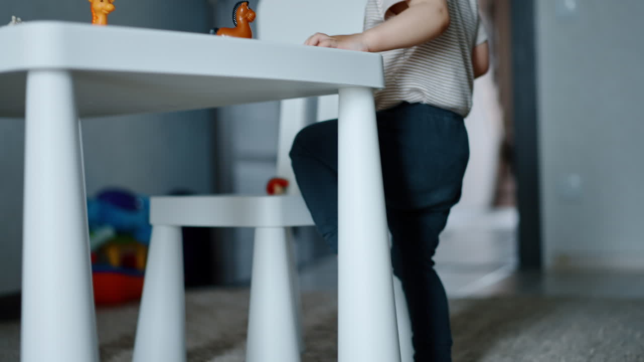 Caucasian child walks barefoot by the fluffy carpet. Boy wants to take a seat by the table but his pacifier is on the chair. Kid throws the pacifier away. Low angle view.