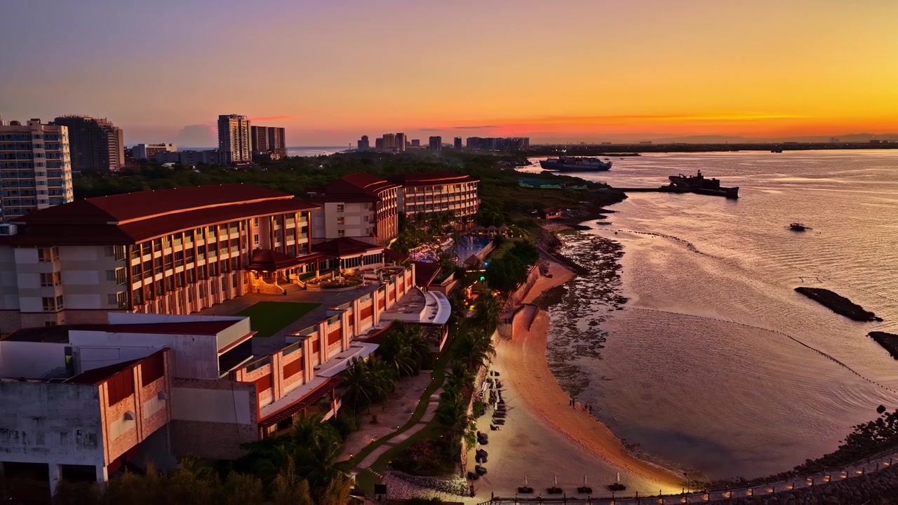 Uno scatto aereo panoramico al tramonto sul resort Dusit Thani Mactan Cebu, che cattura la costa, la spiaggia e gli edifici illuminati dalla luce dorata.