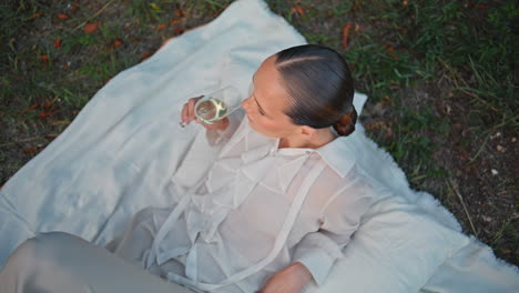 pretty girl tasting wine on white blanket at picnic top view. woman lying grass