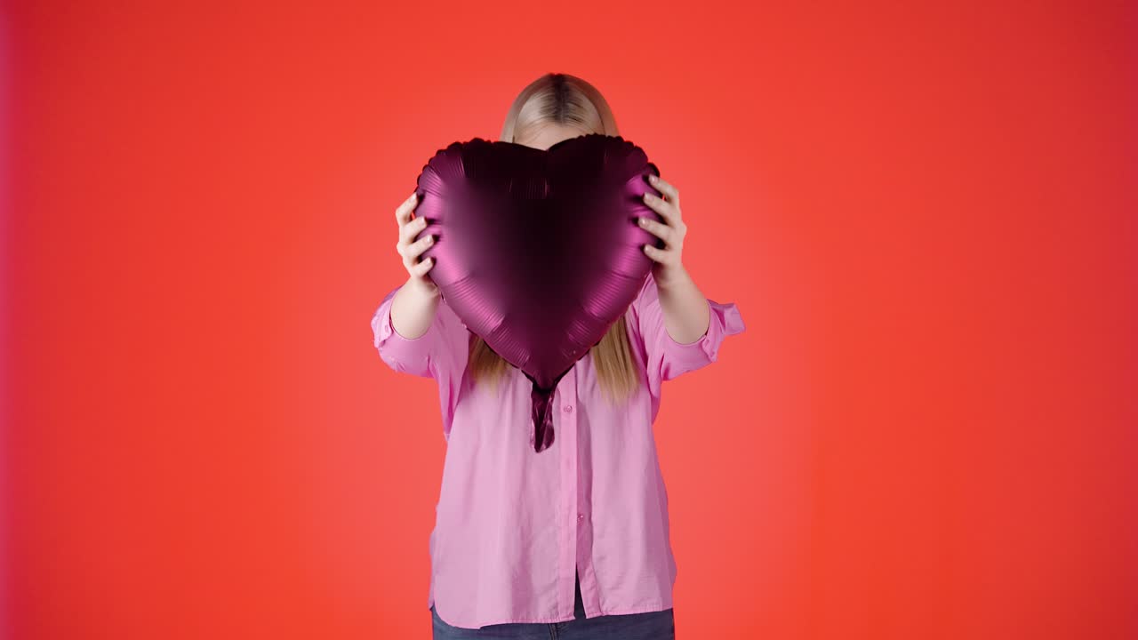 una mujer rubia bonita sosteniendo un globo en forma de corazón púrpura contra un fondo rojo, toma de estudio