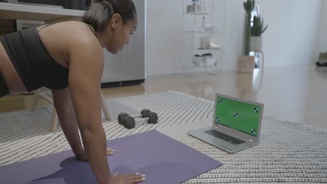 A static shot of a black female doing stretches in front of a green screen on the floor of her house in the kitchen