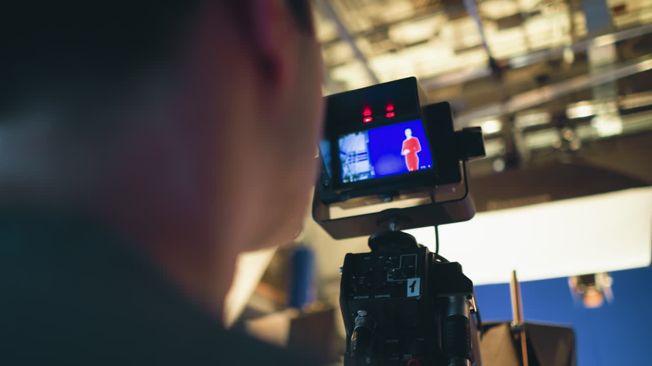 Camera operator monitors screen during live studio shoot with presenter in red outfit on blue screen stage, surrounded by lighting rigs, ceiling mounts, and professional video production equipment in active setup