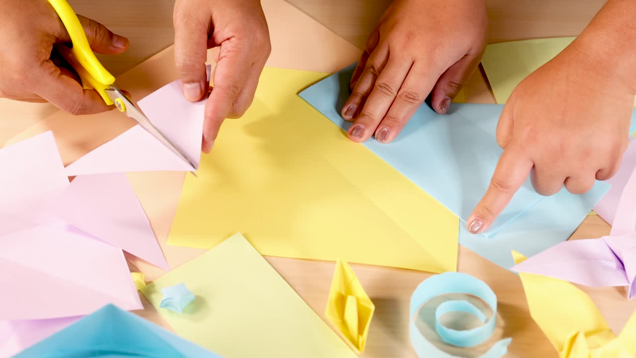 Two people cut and fold pastel origami paper on a table under bright, even lighting