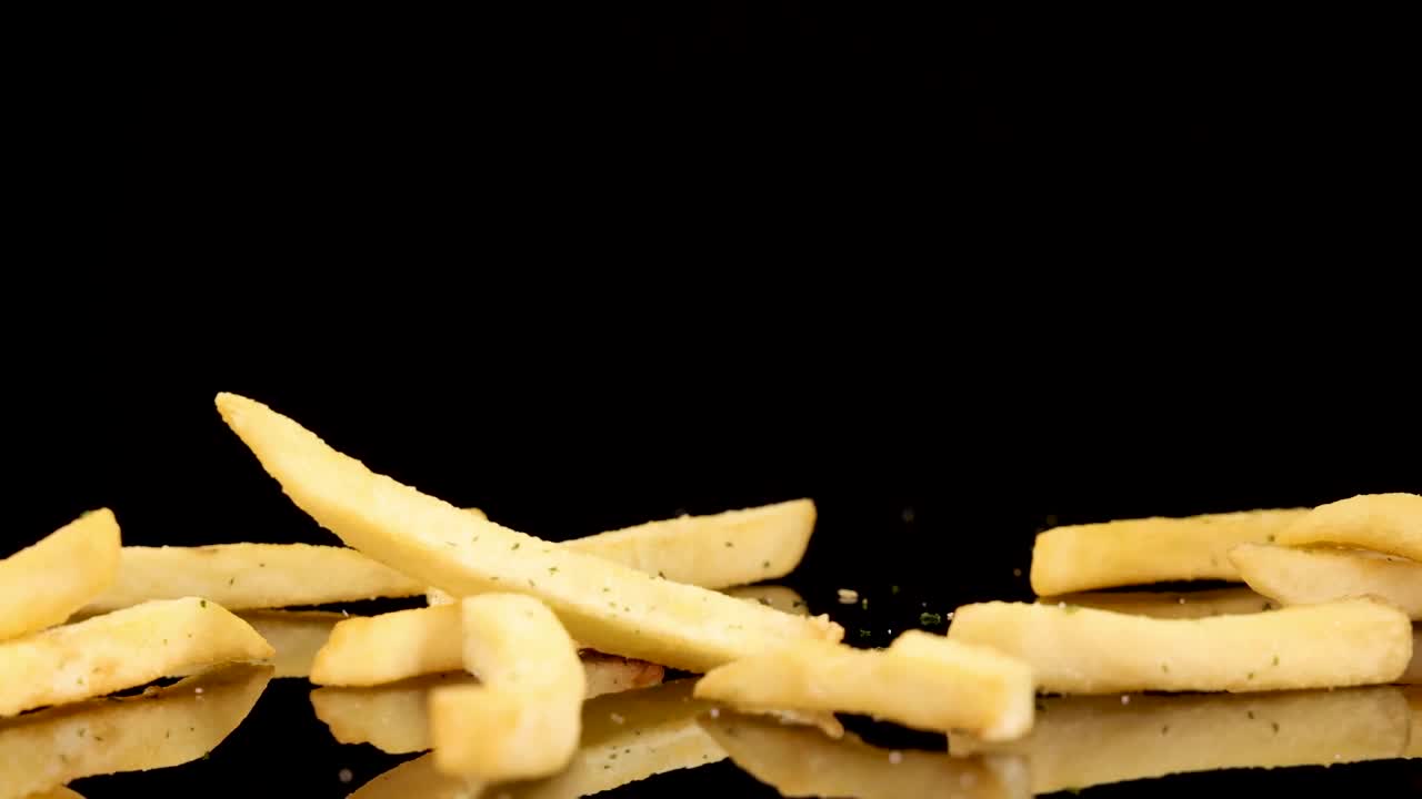 Golden French fries fall and scatter onto a glossy black surface under bright studio lighting, captured in slow motion with a static camera for dramatic effect