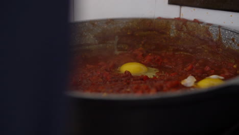 man-breaks-an-egg-into-a-bubbling-tomato-sauce,-preparation-of-homemade-Shakshouka,-filmed-in-slow-motion-and-natural-day-light