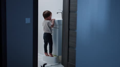 baby boy stands on the stool to reach the sink. curious kid observes how the water flows. black cat enters the bathroom.