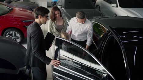 A-family-on-an-excursion-to-a-car-dealership-under-the-supervision-of-a-brunette-man.-An-assistant-in-a-business-suit-a-middle-aged-man-in-a-shirt-together-with-his-son-inspect-the-dealership-and-the-car-from-the-inside