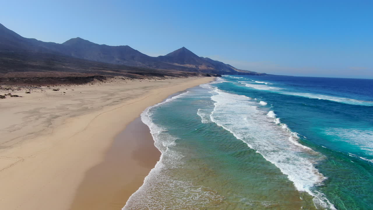 volando sobre la playa de cofete, fuerteventura: vista aérea viajando a la orilla de la gran playa y en el fondo las grandes montañas