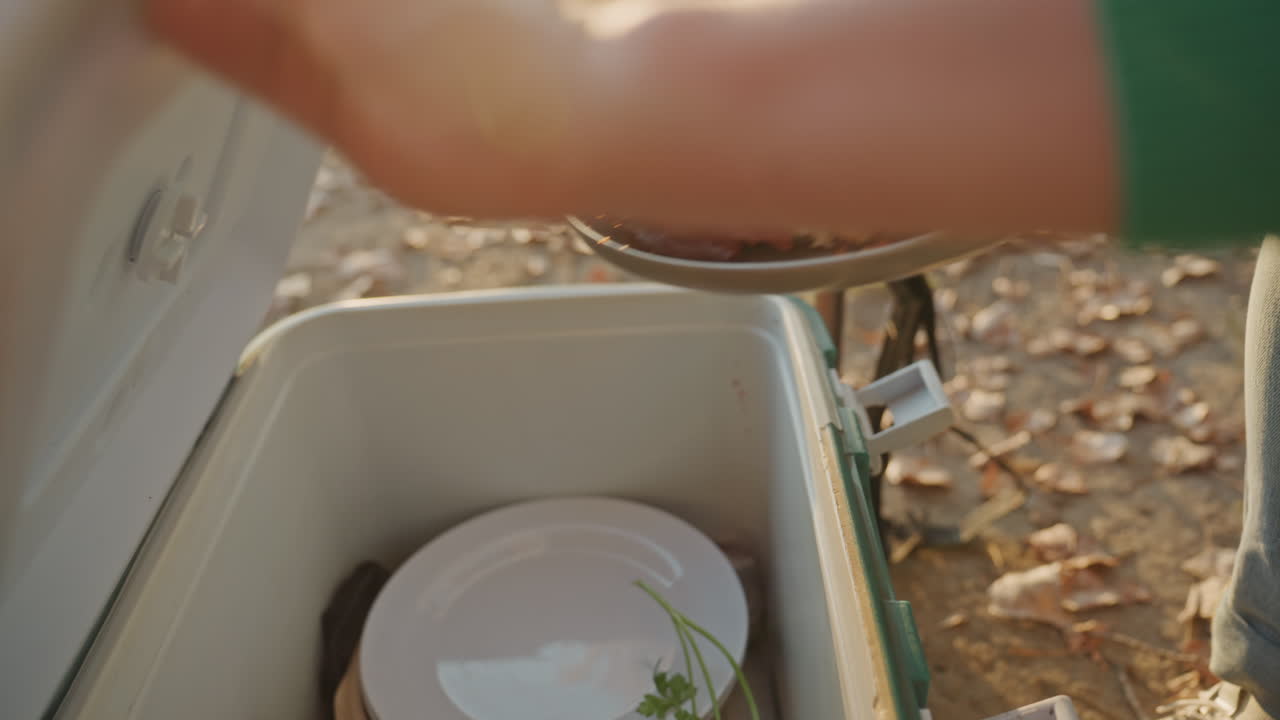 Primo piano della torta sulla borsa frigo sotto la luce del tramonto, la donna solleva una teglia di metallo e poi posiziona la torta nella borsa frigo, il coperchio si chiude accanto al thermos, foglie cadute a terra, calma atmosfera crepuscolare di riordino del campeggio e cibo.