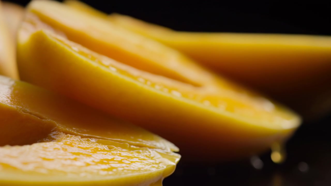 Vista ravvicinata di fette di mango giallo fresco disposte diagonalmente nell'inquadratura con una goccia di succo lucida che pende dal bordo inferiore contro uno sfondo scuro e sfocato.