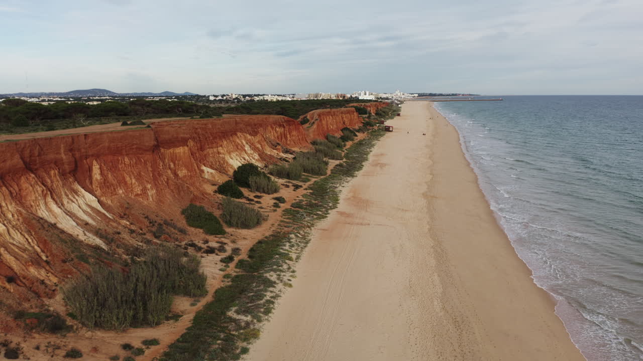 Aerial truck shot of red Sandstone cliffs along sandy beach at Algarve Coastline with resort town in background