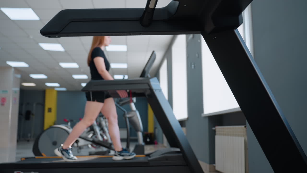close up of treadmill under console showcasing running belt and safety clip while woman in black workout outfit walks in modern gym with exercise bikes and elliptical trainer behind