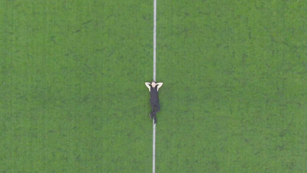 Caucasian man resting, relaxing in the middle of a soccer stadium aerial drone footage. The camera moves higher in the air revealing the whole soccer or football stadium field during summer day time.