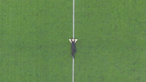 Caucasian man resting, relaxing in the middle of a soccer stadium aerial drone footage. The camera moves higher in the air revealing the whole soccer or football stadium field during summer day time.