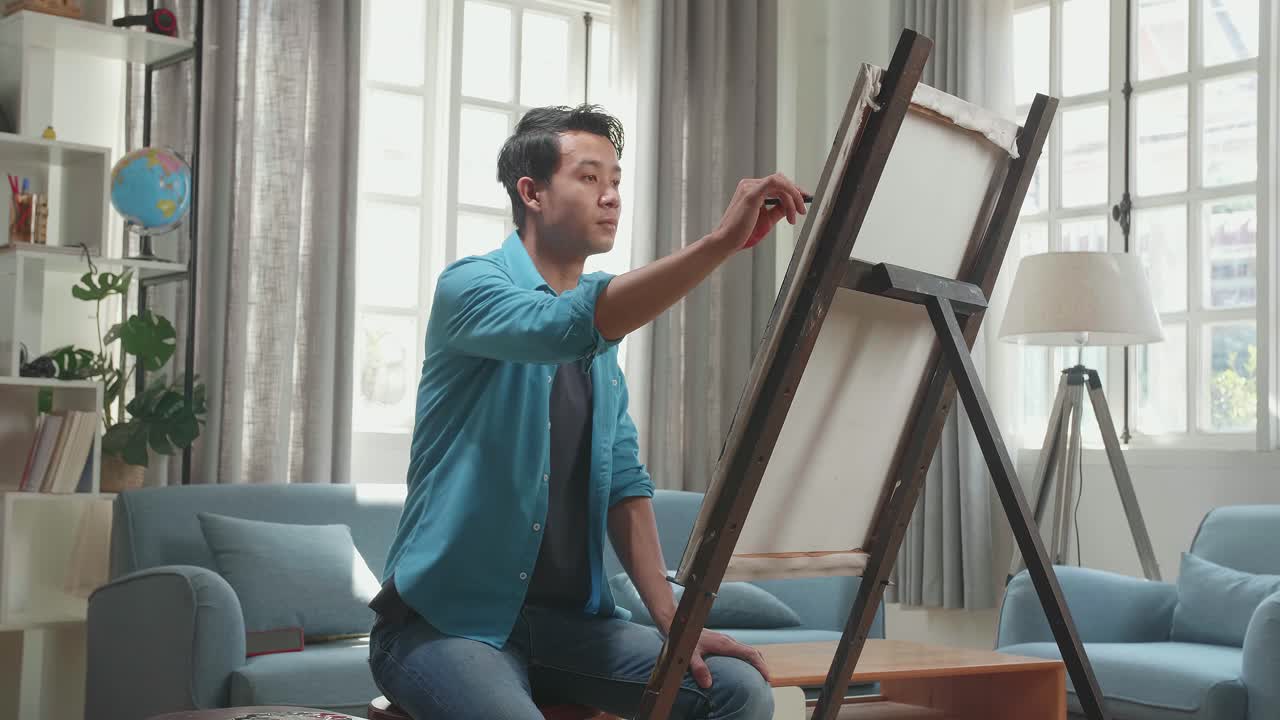 Side View Of An Asian Artist Man Sitting On Round Wooden Chair Without Backrest Holding Paintbrush And Mixing It With Colour Before Painting On The Canvas