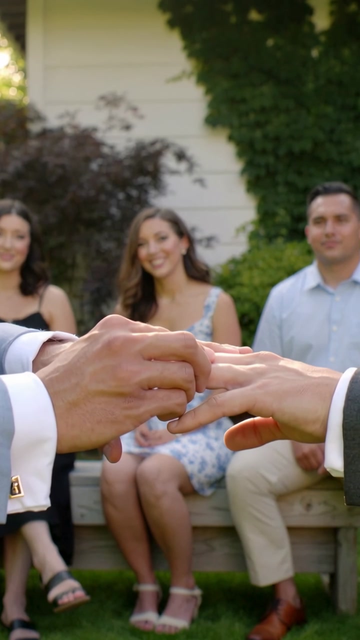 Vertical video: Aligning cufflinked hand sliding ring onto left ring finger to wed in yard, guests