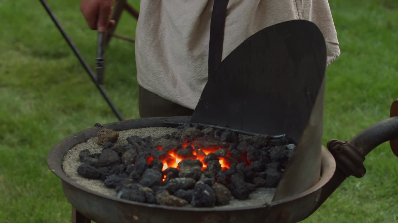 un herrero reinacter de la guerra revolucionaria pone una pieza de metal en brasas para calentar mientras bombea el fuelle