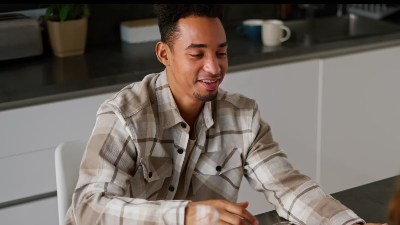 Close up of a happy young man with Black skin color in a checkered beige shirt drinks orange juice with his girlfriend during his breakfast in the kitchen