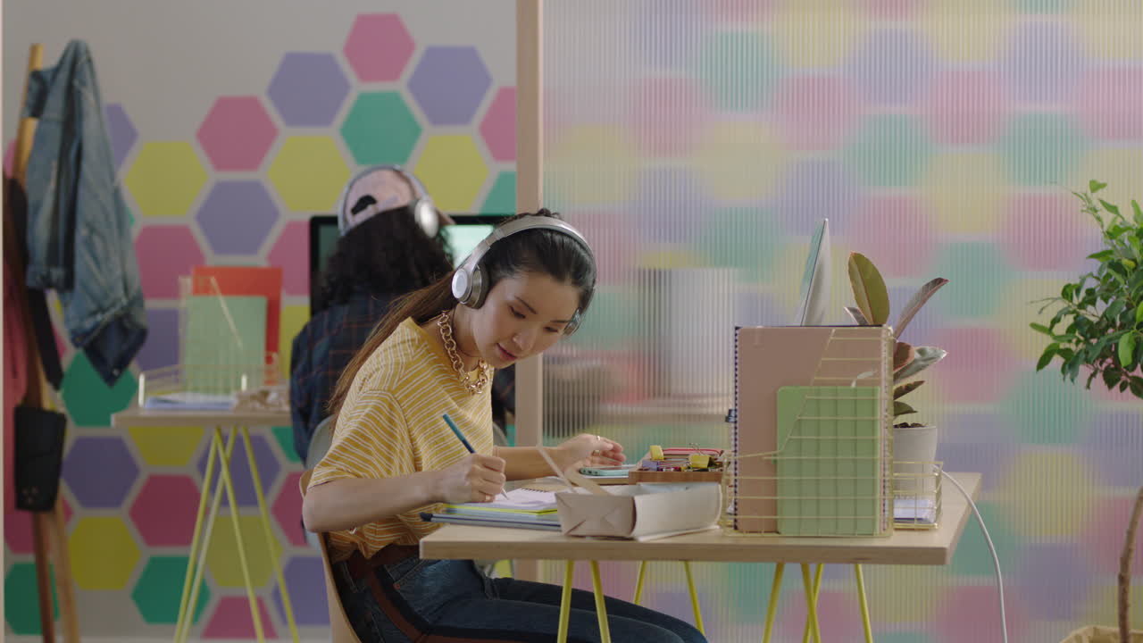 hermosa joven asiática estudiante usando computadora brainstorming trabajando en un proyecto creativo disfrutando del estudio en una oficina moderna colorida escuchando música usando auriculares