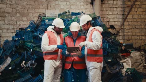 Ein-Trio-Von-Arbeitern-In-Weißen-Uniformen-Und-Orangefarbenen-Westen-Arbeitet-An-Einem-Laptop-Und-Kommuniziert,-Während-Sie-Neben-Einem-Berg-Aus-Plastikmüll-In-Einer-Müllrecyclinganlage-Stehen.