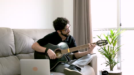 bearded hispanic man playing acoustic guitar and singing on sofa in front of laptop