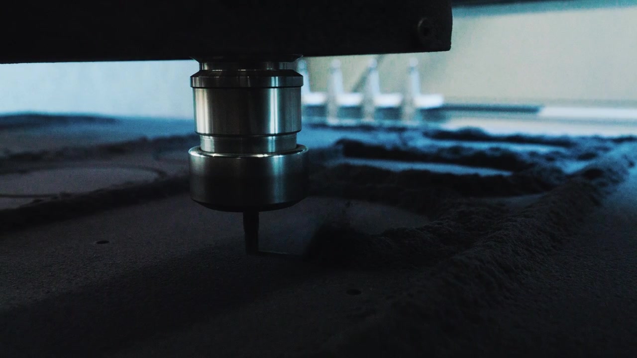 Primo piano dal basso di una fresa CNC che taglia una tavola scura con polvere sottile lungo la superficie, al rallentatore.