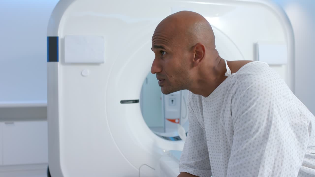 Uomo afroamericano che si prepara alla tomografia computerizzata indossando un camice, appoggiato al tavolo, che guarda il monitor, spazio per la copia