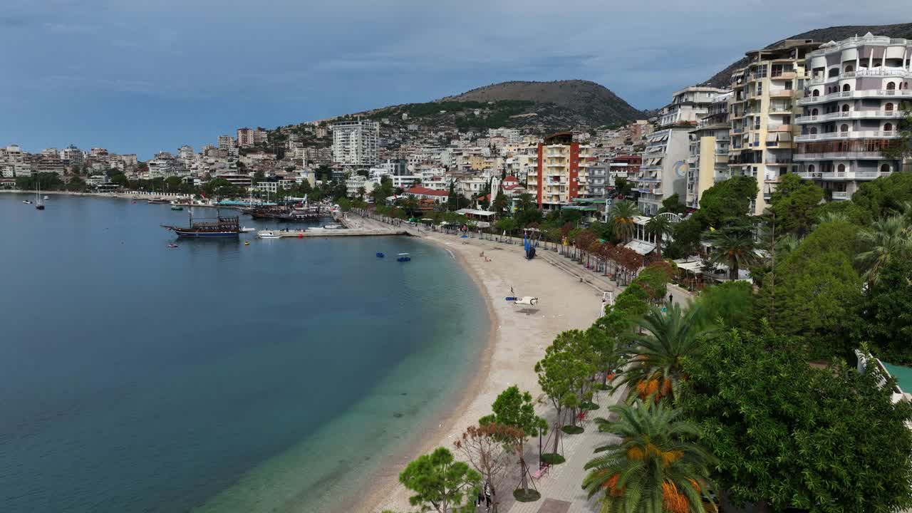 Saranda coastline with white sand beach, calm turquoise waters, anchored boats, and layered buildings rising into the hillside, Albania