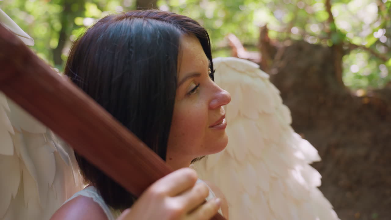 Angel woman with white wings holding wooden staff, eyes closed in gentle forest light, expressing deep peace and harmony with nature, serene face illuminated by warm sunlight
