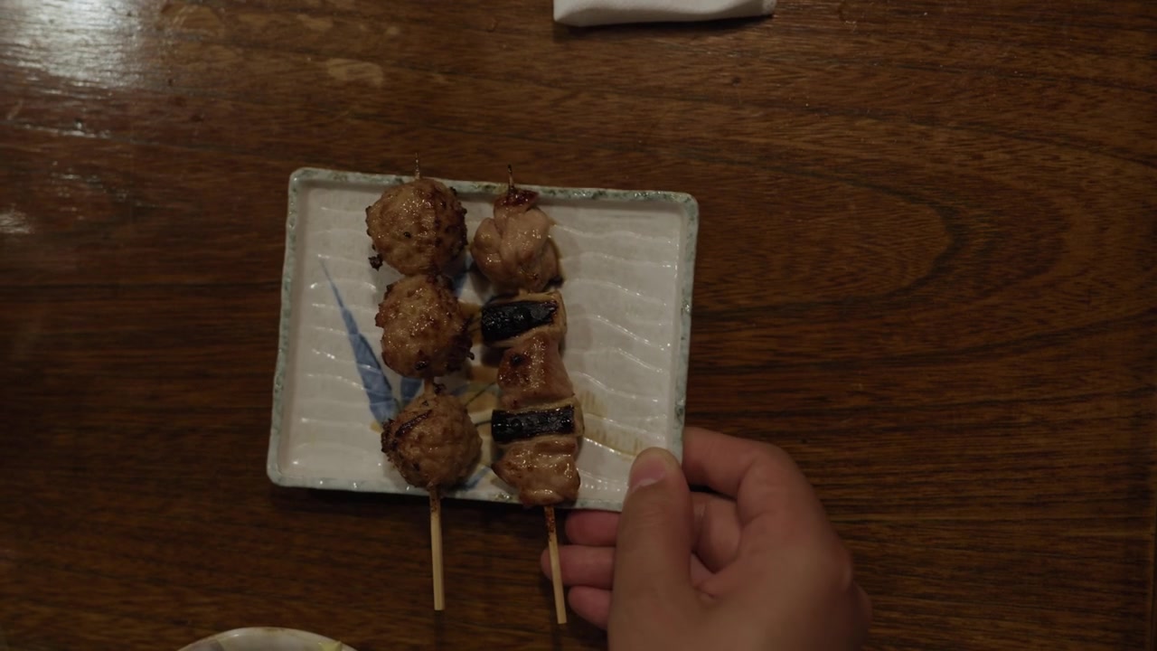 Hand placing a ceramic plate of grilled yakitori chicken skewers and tsukune meatballs on a wooden table in Japan