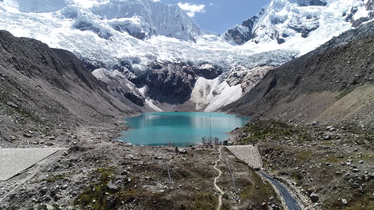 A stunning turquoise glacial lake sits beneath snow-capped peaks, with visible infrastructure