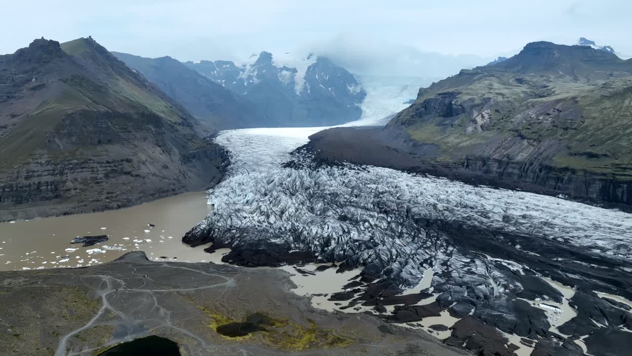 Luftfoto af Vatnajökull-gletsjeren i Island, der viser dens isbræ, som løber ud i en mudret smeltevandssø mellem stejle vulkanbjerge