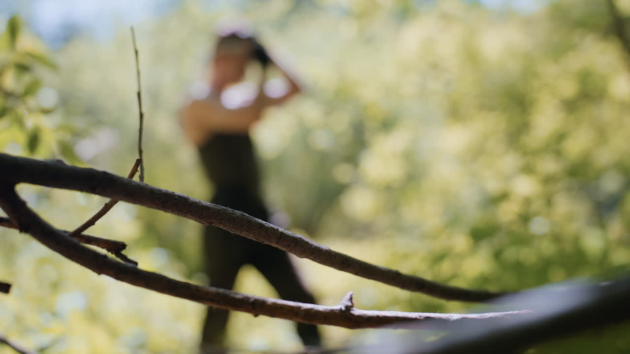 Sharp focus on rugged tree branch in foreground with blurred adventurer adjusting headband under sunlit forest canopy, creating depth and serene outdoor adventure atmosphere on summer trek