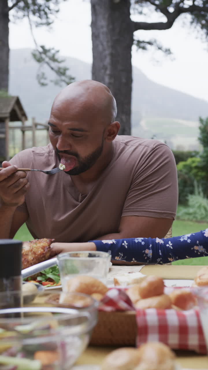 Vertical video of biracial father and daughter eating meal at dinner table in garden, slow motion