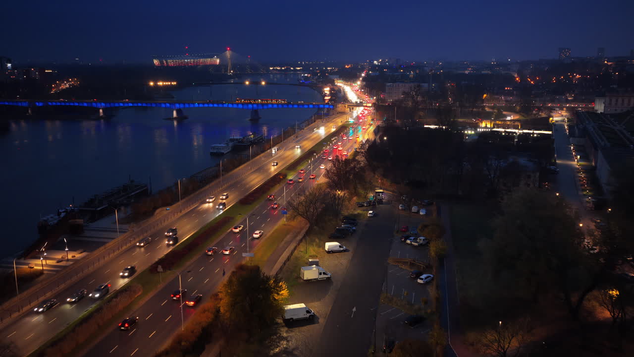 Aerial drone view of Wybrzeze Gdanskie road along the Vistula River, showing heavy evening traffic, city lights, and the illuminated Swietokrzyski Bridge and National Stadium