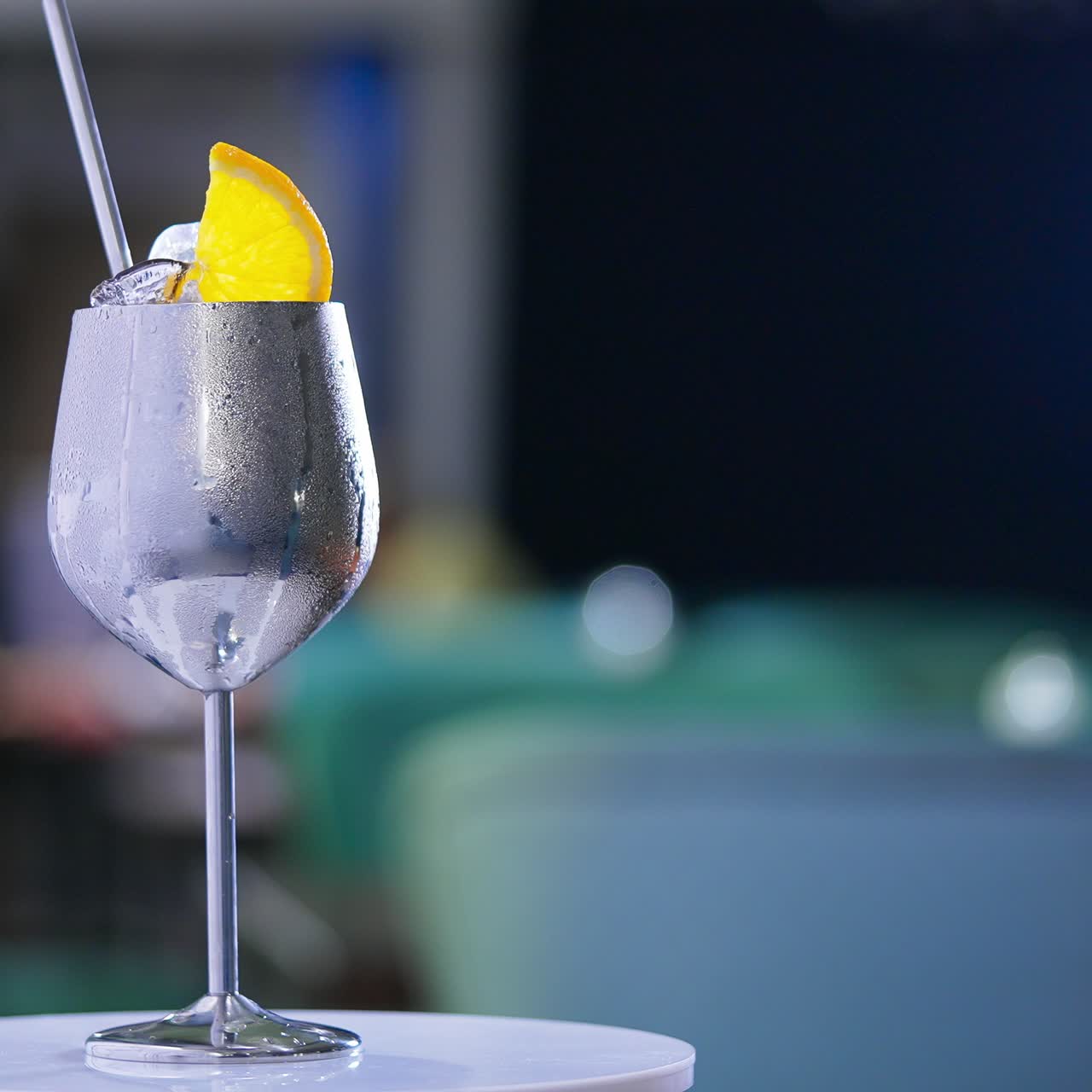 Misted metal wineglass with metal straw inside on the rotating table. Cocktail with ice cubes and orange slice on top. Close up. Blurred backdrop