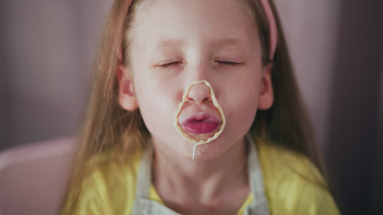 Close up of cheerful girl wearing yellow shirt playfully adjusting cutout dough around her mouth during fun creative baking session, showing humor and imagination in kitchen playtime