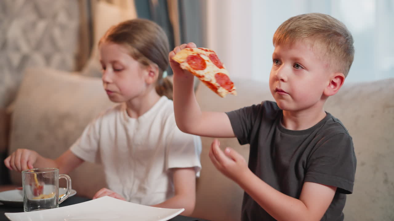 boy enjoys cheesy pepperoni pizza while girl dips fries into sauce as they sit together on couch focused on something off screen, sharing casual meal in cozy home setting