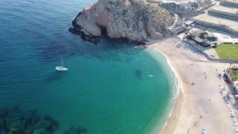 Aerial-panning-shot-of-the-wonderful-medano-beach-in-cabo-san-lucas-overlooking-the-blue-sea-with-boats-in-the-water,-hotel-buildings-and-majestic-mountains-in-the-background-on-a-sunny-day